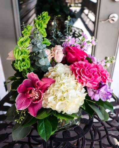 Hot Pink Party Spring bouquet featuring hot pink roses, white hydrangeas, purple orchids, cream spray roses, lavender stock, and bells of Ireland, all arranged in a clear glass cube vase
