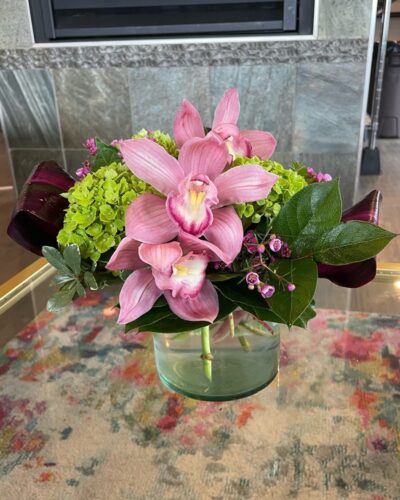 "Paradise Found" flower arrangement with pink orchids, green hydrangeas, pink waxflower, burgundy ti leaves in a clear glass vase.