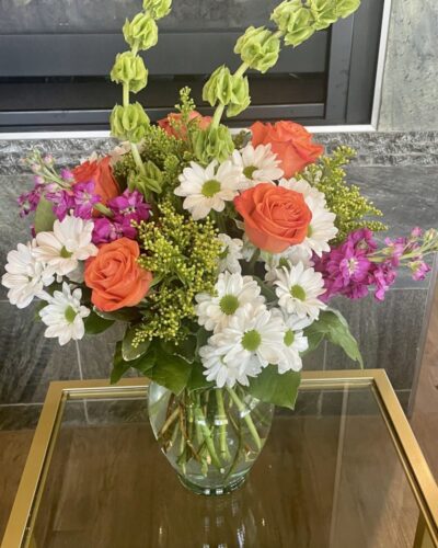 "Lucky to Love You" bouquet with orange roses, white daisies, green carnations, bells of Ireland in a clear vase.